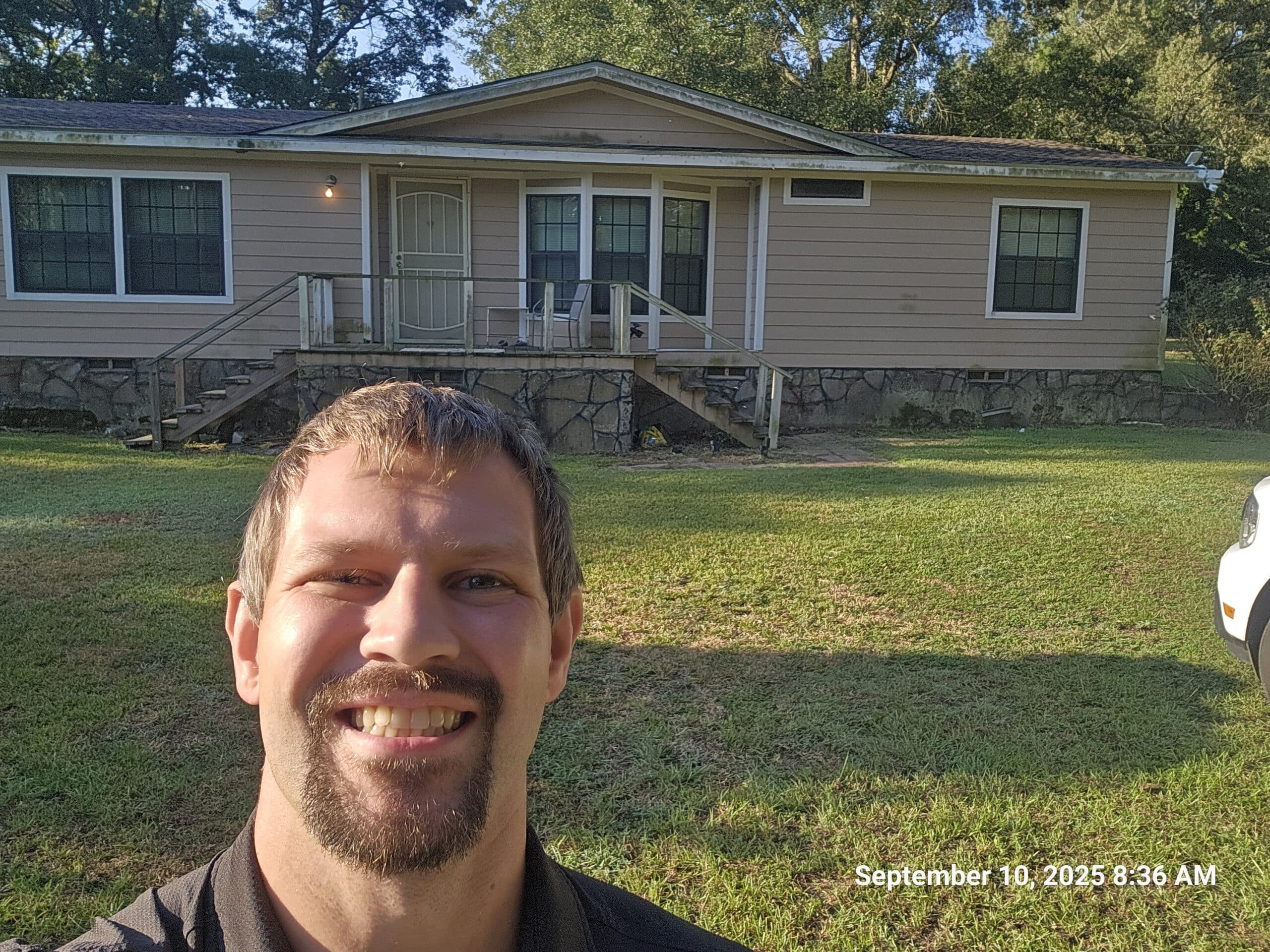 Mobile home with elevated front porch and stone skirting on a grassy lot, with an inspector standing in the foreground during a foundation evaluation.